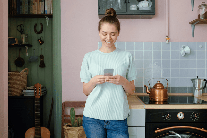 Young smiling woman holding her smart phone with both hands, messaging online, communicating with friends, checking newsfeed on social networks, standing in nice pastel kitchen