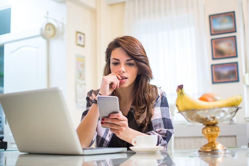 Young woman sits at a table in front of her computer, a phone and a bowl of bananas, all while biting her nails. Biting nails, along with some other habits can hurt your jaw - leading to TMJ disorders.