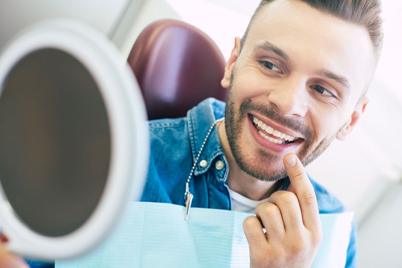 a cosmetic dentistry patient smiling and pointing at his enhanced smile in a hand held mirror after getting treated with teeth whitening.